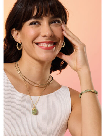 A head and shoulders photo of a model wearing gold coloured jewellery, including a Gold-coloured gemstone necklace with large prehnite and feather pendant