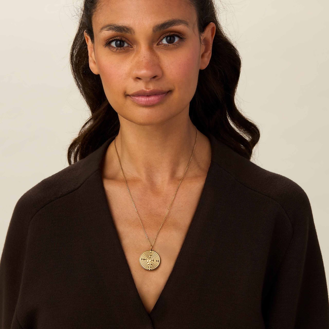 A woman in a brown, v-neck top, wearing a gold chain necklace with a labyrinth of life pendant from ENERGETIX
