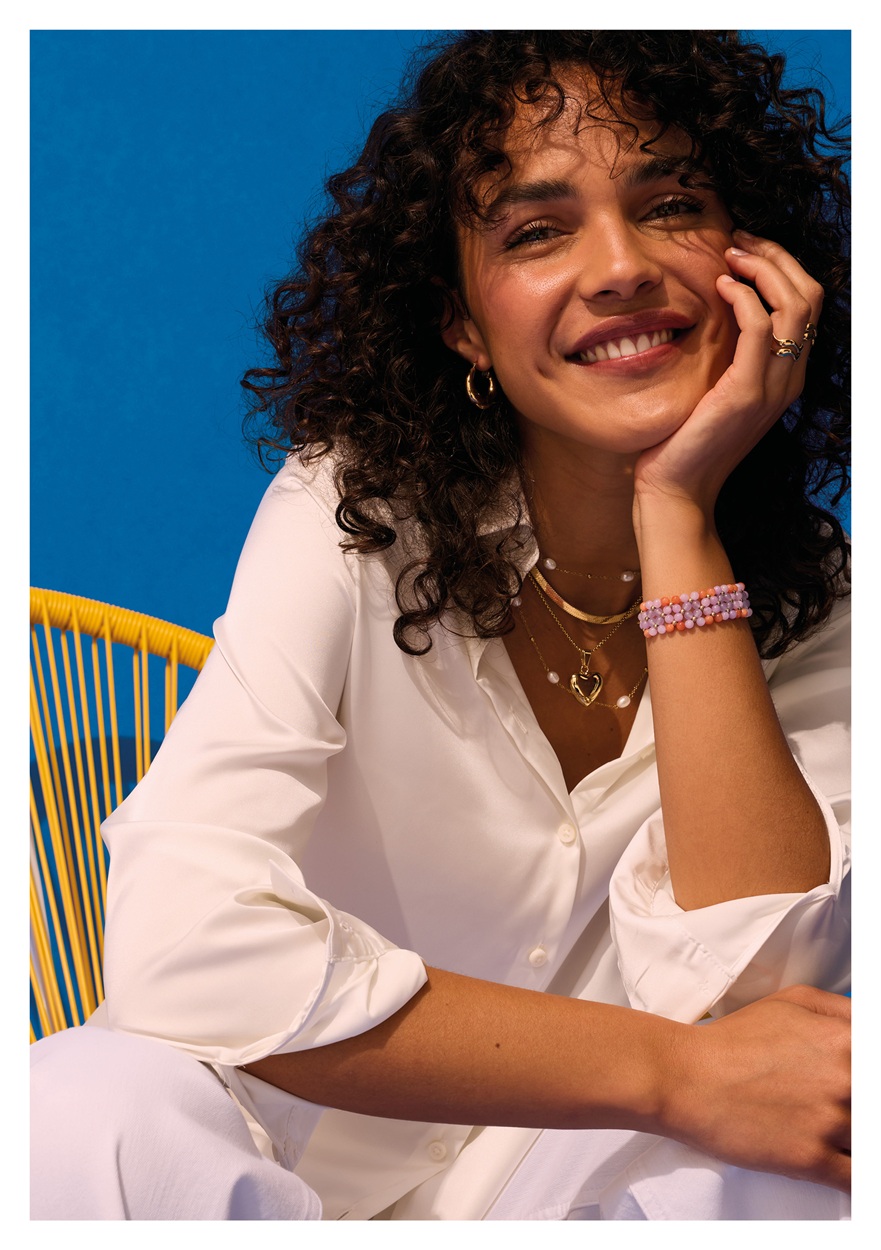 A model sitting in front of a bright blue sky, smiling directly at you. She is wearing a collection of ENERGETIX Magnetic Jewellery