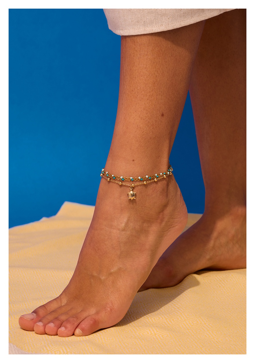 A close-up of a model standing on sand, wearing a couple of gold coloured ENERGETIX ankle bracelets, one with a turtle pendant.